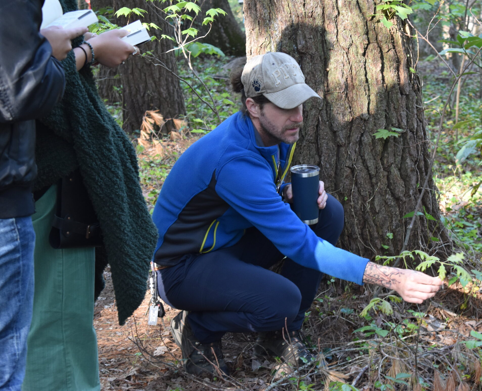Professor Eric Griffin squats beside a tree and shows a group of students leaf structures.