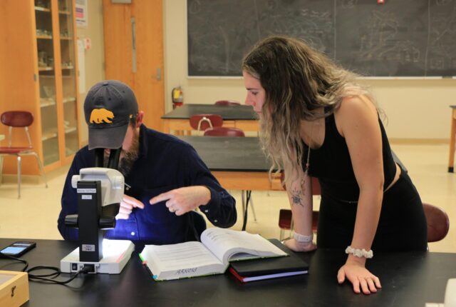 A biology and environmental studies crew student helps another student study a sample through a microscope