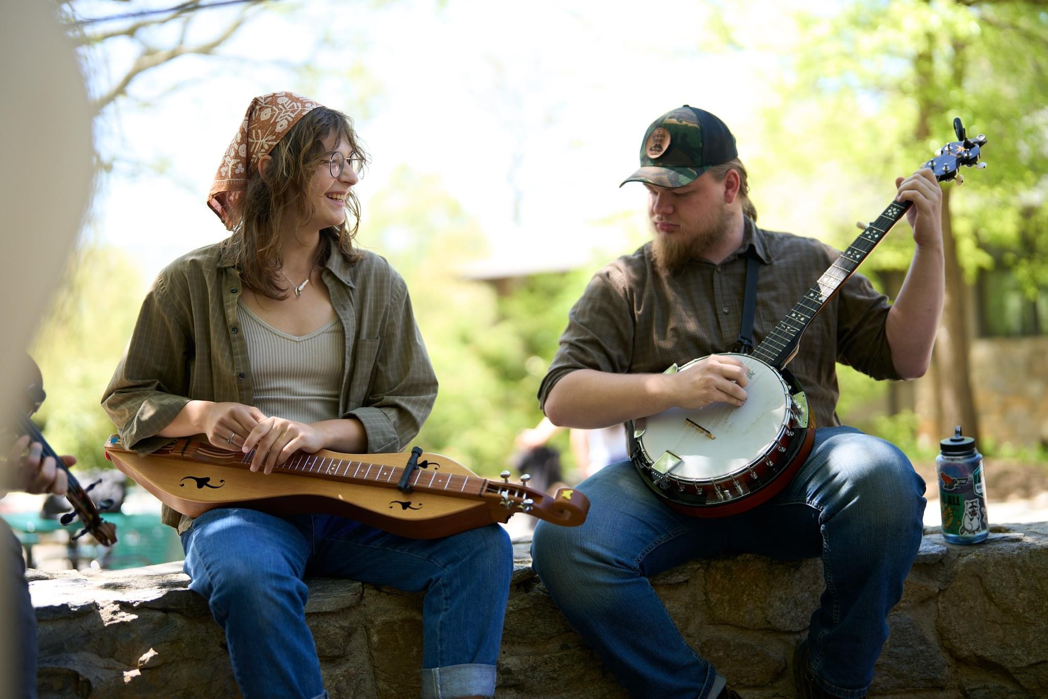 Two students sit on a rock wall with a banjo and dulcimer in hands smiling.