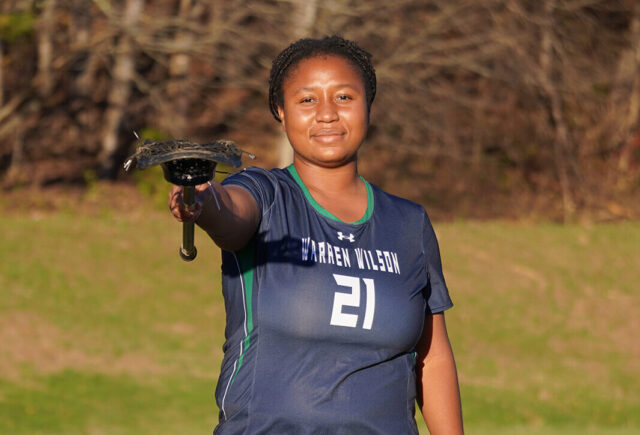 Madison Sings, a senior at Warren Wilson College, stands in her lacrosse jersey and points her lacrosse stick to the camera.