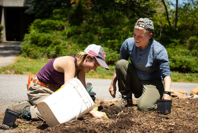 Two Students smile and laugh as they weed a flower bed.