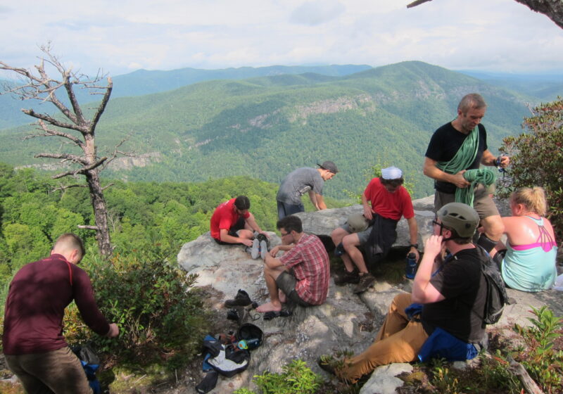 Students on top of a cliff relaxing with the view of nothing but mountains and sky behind them.