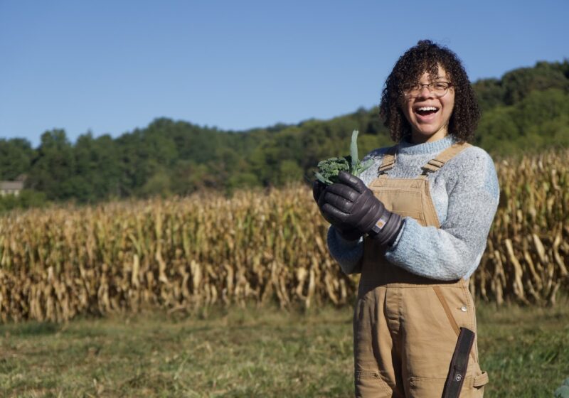 A student is smiling holding a head of broccoli standing in a field of corn.