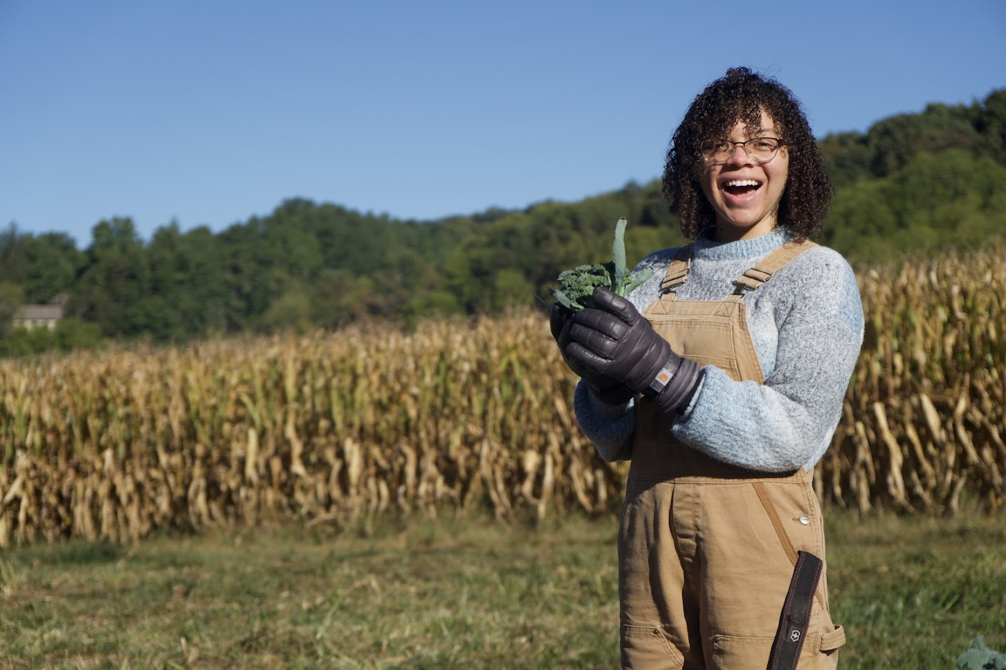 A student is smiling holding a head of broccoli standing in a field of corn.
