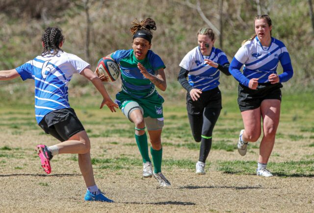 Four people are running in a rugby game. Three members of the opposing team are surrounding the one Warren Wilson Team member holding the ball.