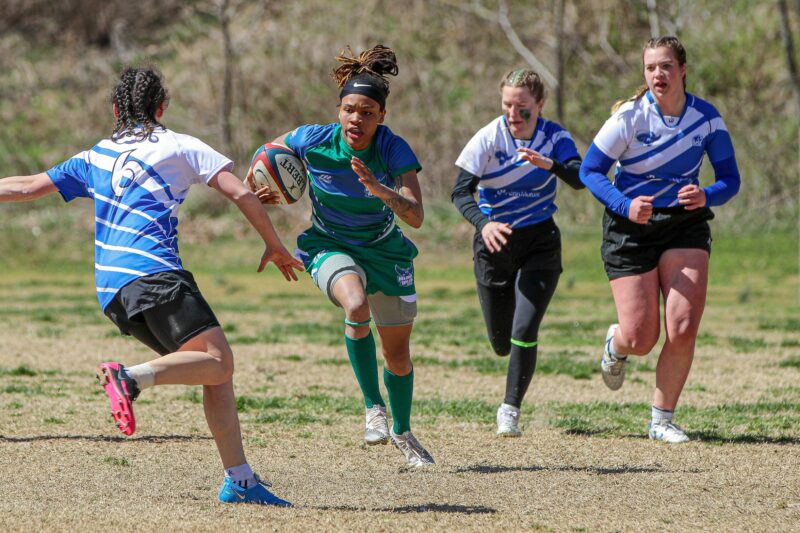 Four people are running in a rugby game. Three members of the opposing team are surrounding the one Warren Wilson Team member holding the ball.