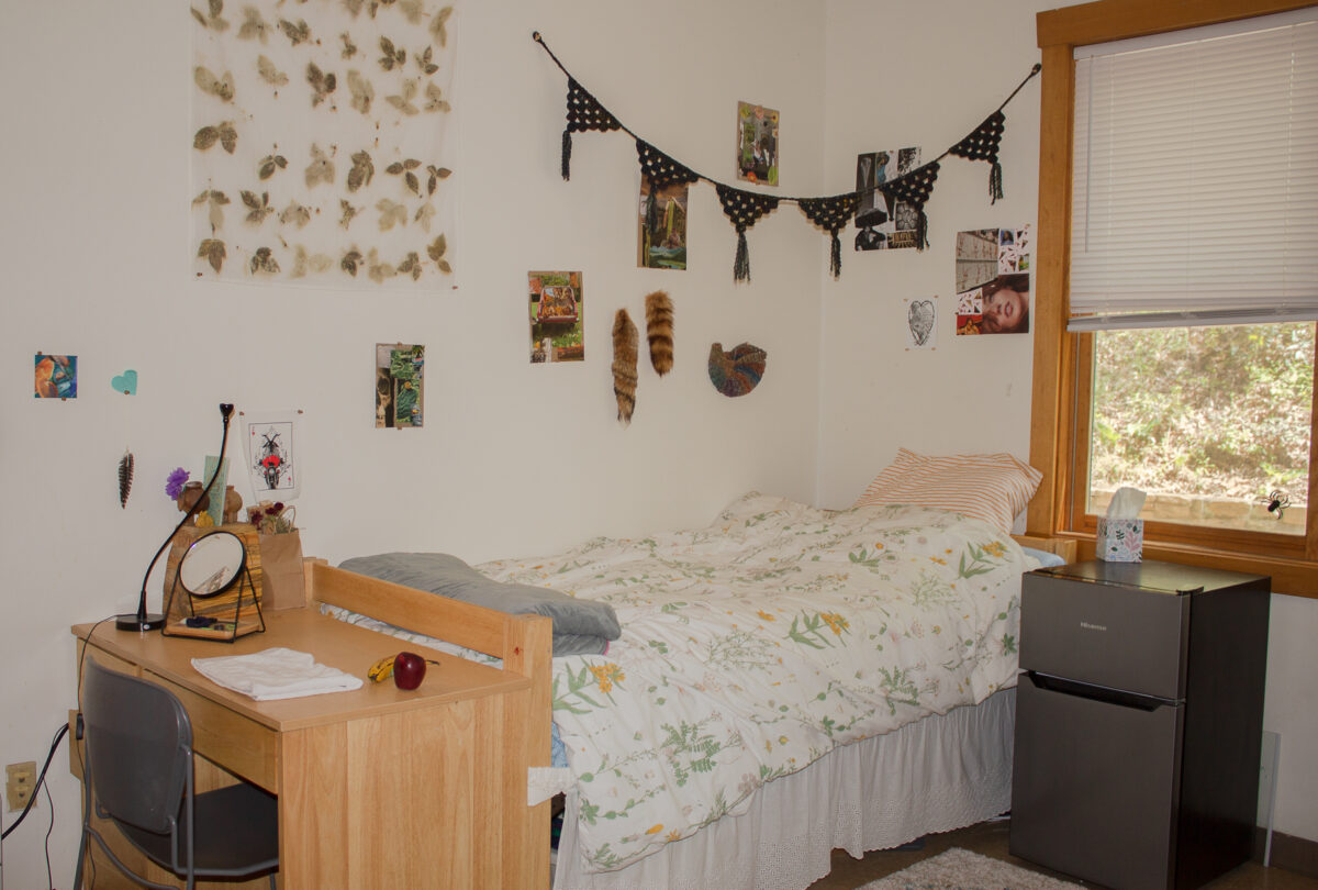 A Wellness dorm room. There is a bed with a desk at the end and a miniature fridge beside it. The walls have picture and decorations hung up.