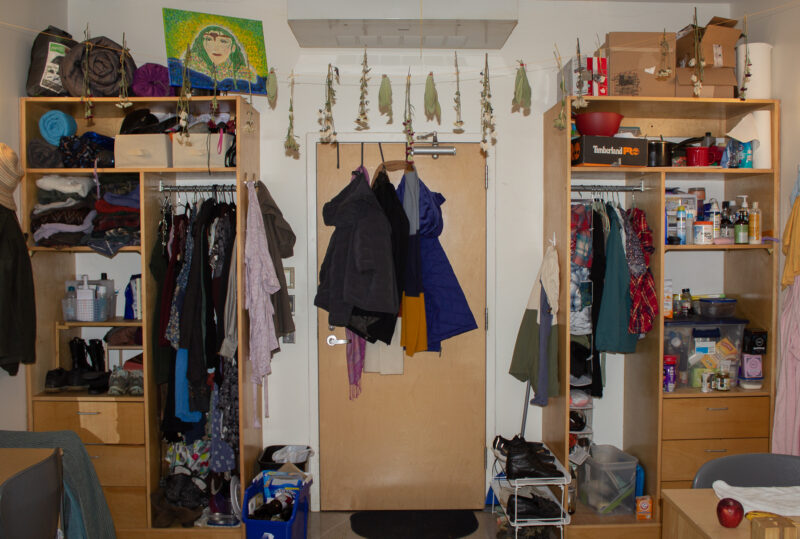 A dorm room in the Wellness Dorm. There are two wooden armoires with clothes and shoes. There are wildflowers and plants hanging from a string spanning the full width of the room. There are coats and scarfs hanging from the back of the door.
