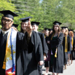 Students walk in the processional. One student looks at the camera and holds out a peace sign