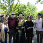 A graduate, surrounded by a group of their family and friends, poses with their degree and pine tree smiling.