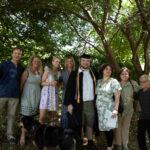 A large group and a dog surround a graduate. They are all smiling