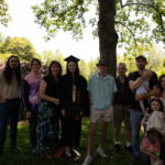 A large group surrounds a graduate. The grad is holding their degree