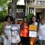 A graduate is joined by four people in a photo. They are all smiling. The Graduate is showing their degree.