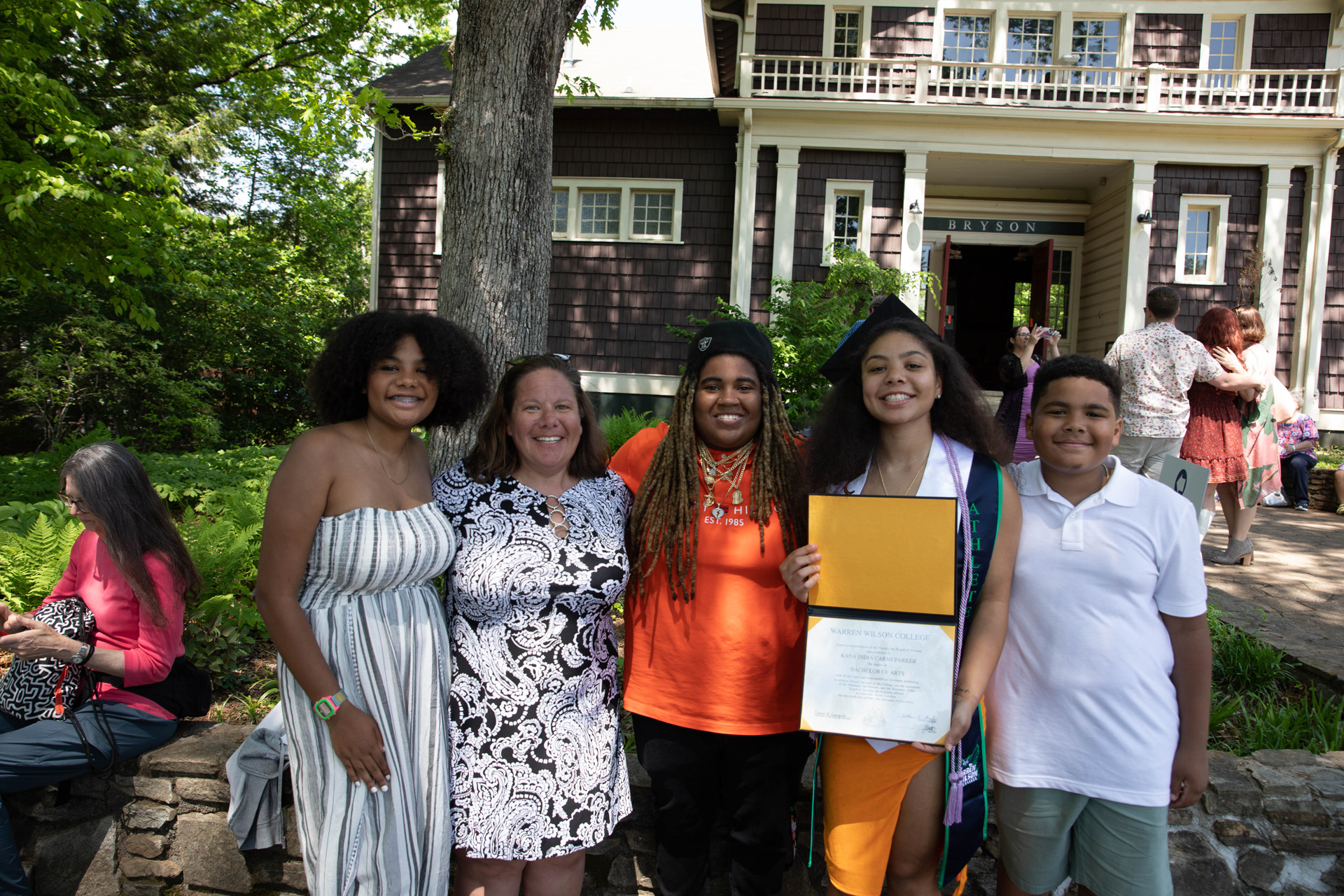 A graduate is joined by four people in a photo. They are all smiling. The Graduate is showing their degree.