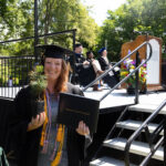 A graduate stands smiling with their tree and degree