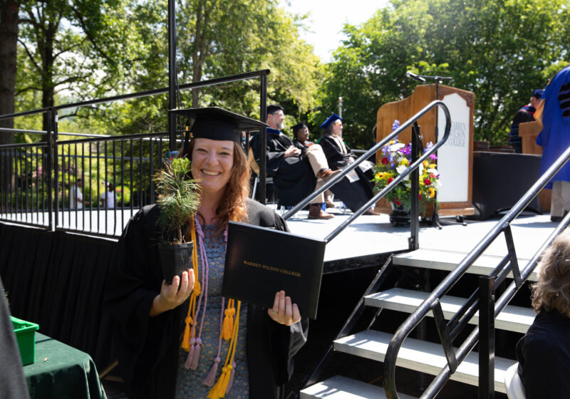 A graduate stands smiling with their tree and degree