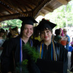 Two grads stand holding their degrees and trees