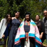 A graduate wrapped in a ceremonial blanket is surrounded by their family and friends. They are holding their degree.