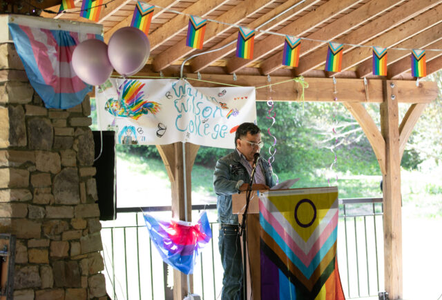 Director of the Queer Resource Center, Jo Go, speaks to those gathered at the lavender graduation. Various Pride flags are hung up in the background.