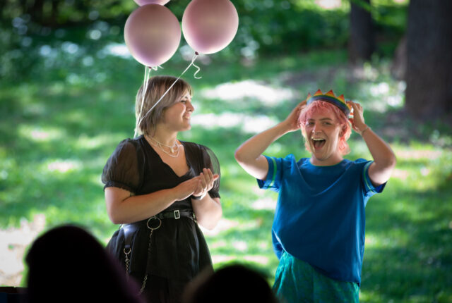 A student wearing a pride crown smiles widely while another student claps for them.