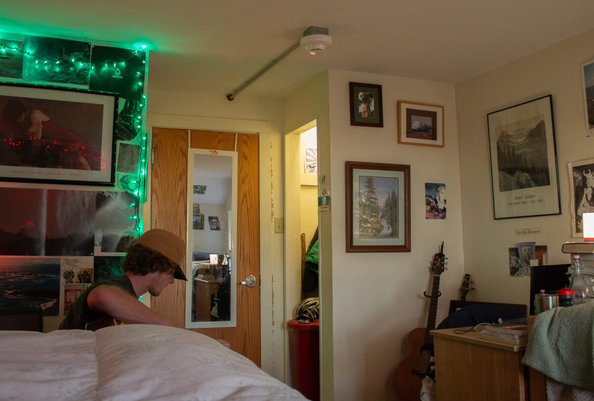 A student plays their guitar in their dorm room. The walls are covered in pictures and lights.