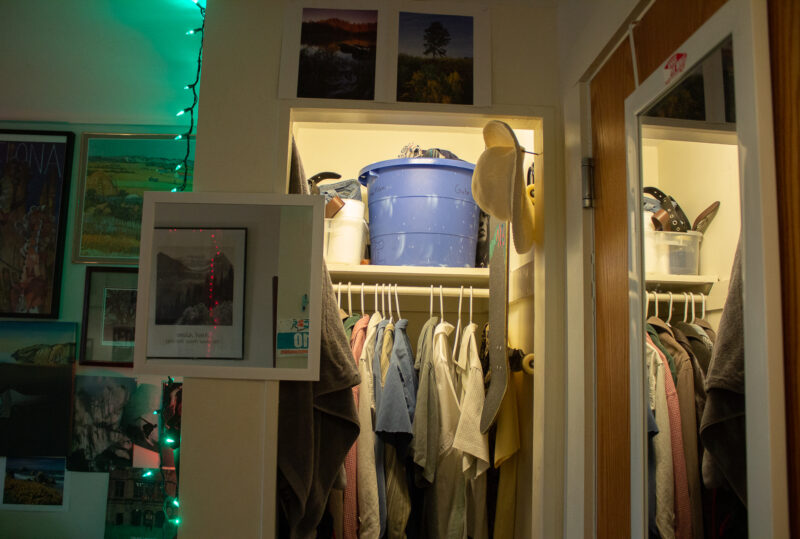 A closet in Sage Dorm. Clothes are neatly hanging, there is a top shelf being used for storage, and a skateboard hangs on the wall.