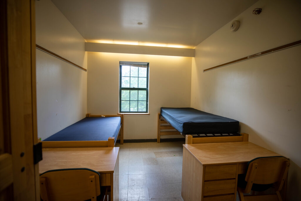 A Dorm room inside Sunderland. There are two twin-sized beds, two desks seen in this photo. There is a window at the far end of the room.