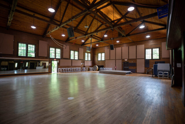 Bryson Gym has a wooden floor. There is a rockwall on the left side of the building and a stage on the right. There is a large mirror that spans nearly the entire rear wall of the gym.