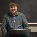 Communications Professor Beck Banks sits at a desk with a macbook smiling