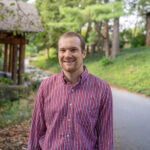 Faculty member Christopher Potvin stands smiling in a red shirt in front of Orr Cottage