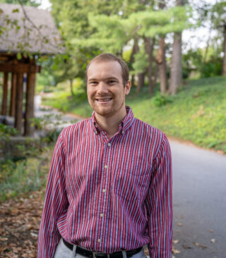 Faculty member Christopher Potvin stands smiling in a red shirt in front of Orr Cottage