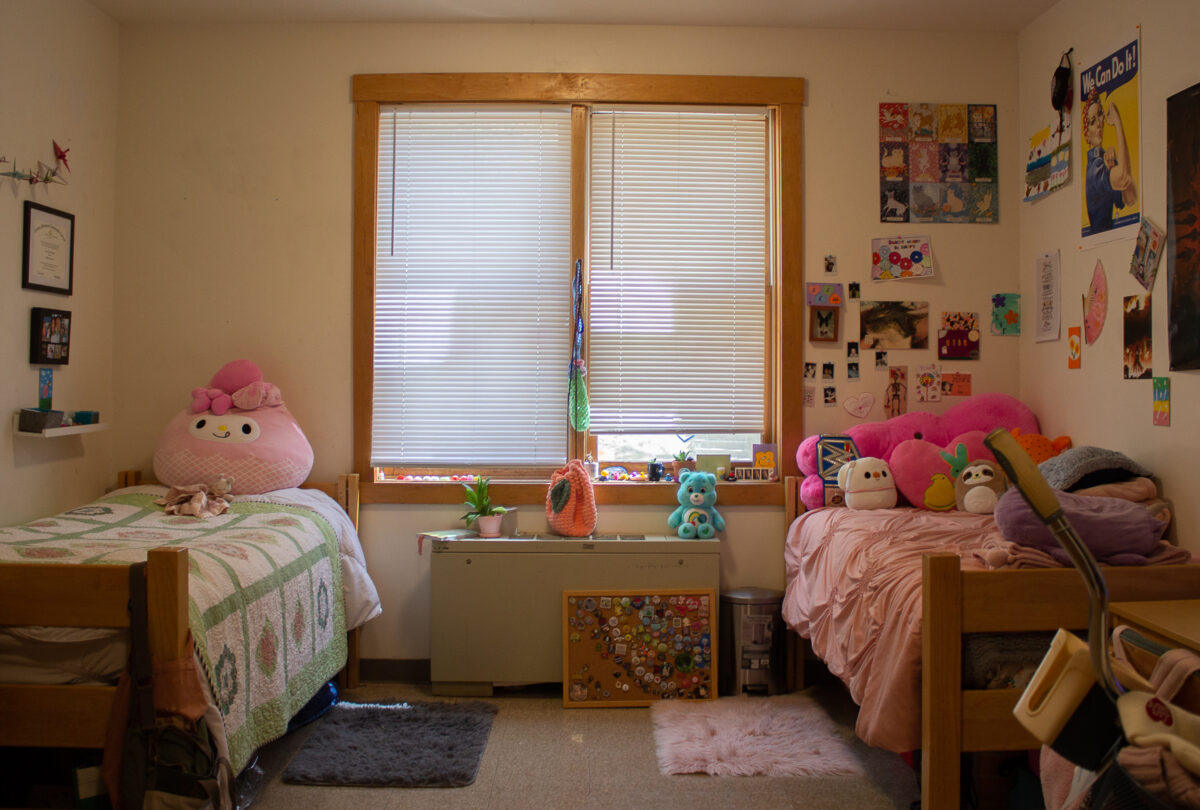 A dorm room in antc with two beds in the focus. one side of the room is covered in posters and pictures.