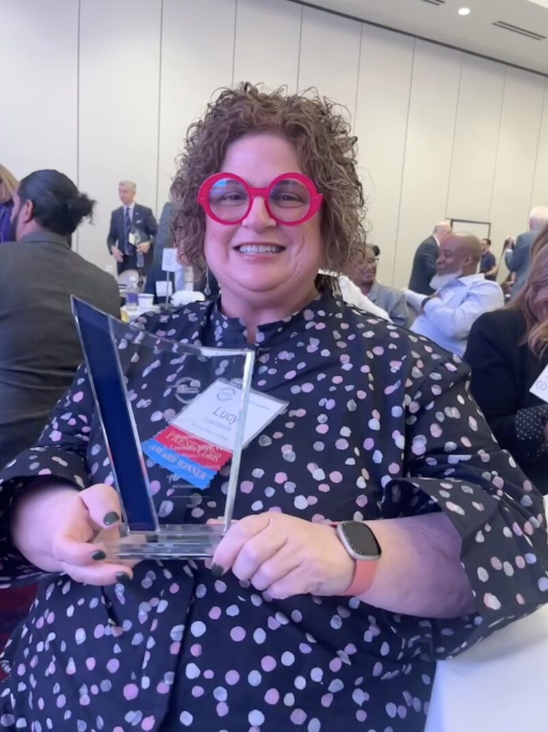 Dr. Lucy Lawrence stands at the PACE Conference holding the Engaged Faculty Award. She is wearing bright red glasses and a polka dot shirt.