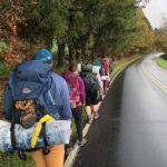 A group of students backpacking walk on the side of a road