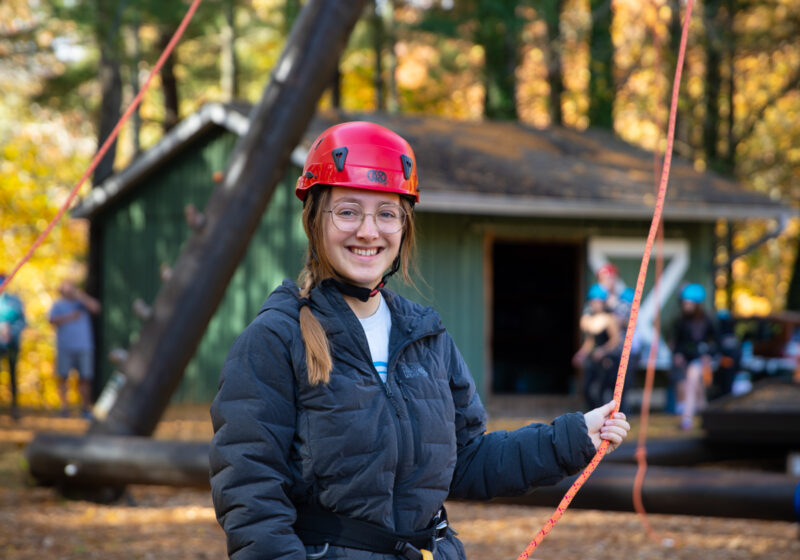 Student Ahna Webster stands at the base of the Alpine Tower challenge course holding ropes and smiling.