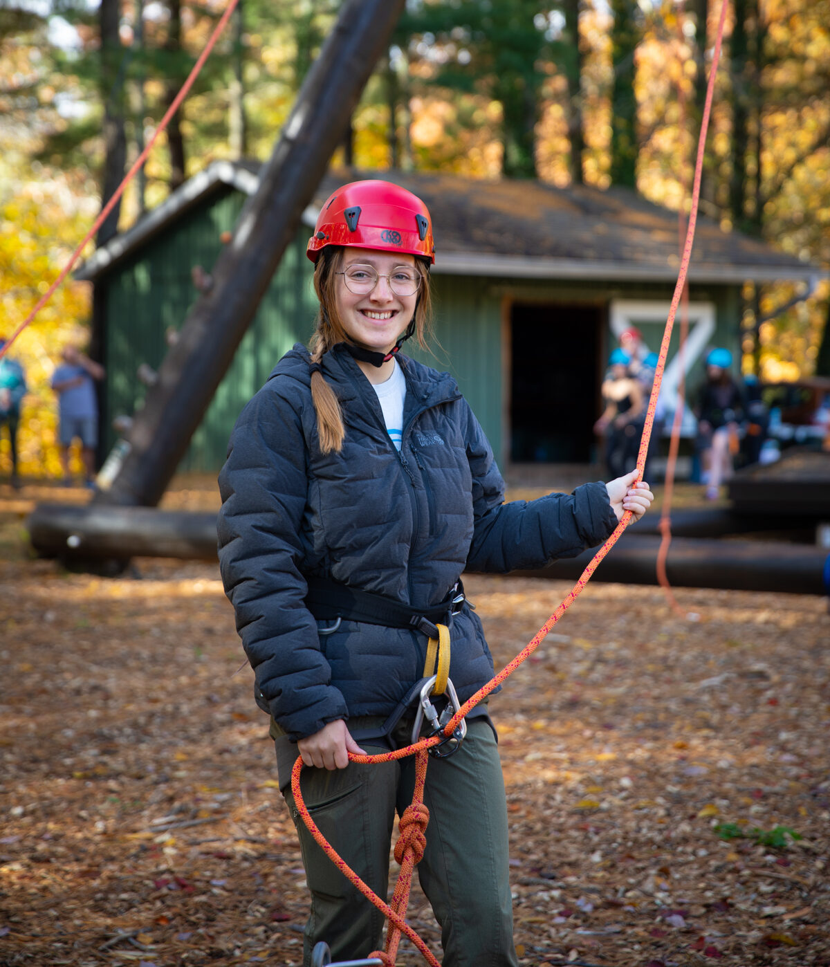 Student Ahna Webster stands at the base of the Alpine Tower challenge course holding ropes and smiling.