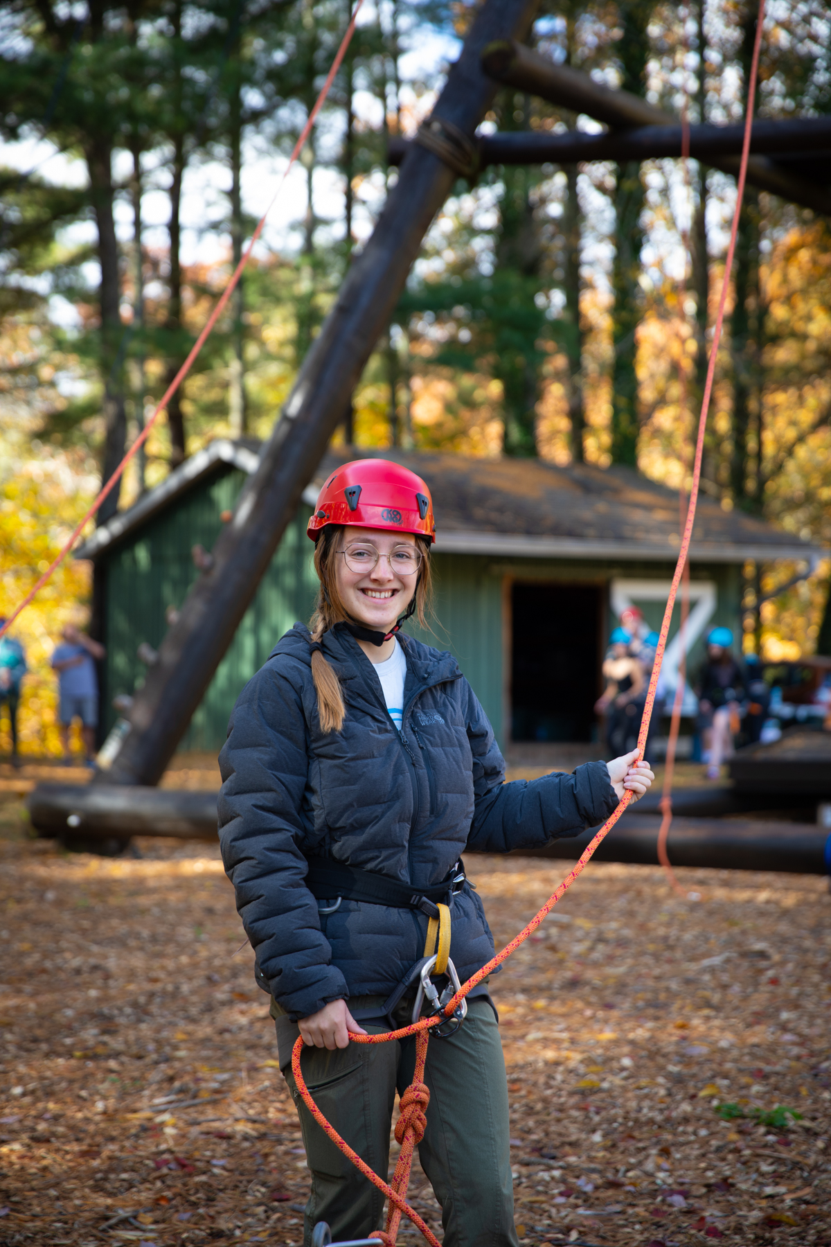 Student Ahna Webster stands at the base of the Alpine Tower challenge course holding ropes and smiling.