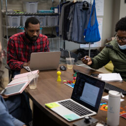 Students sit at a table in front of computers and discuss a project