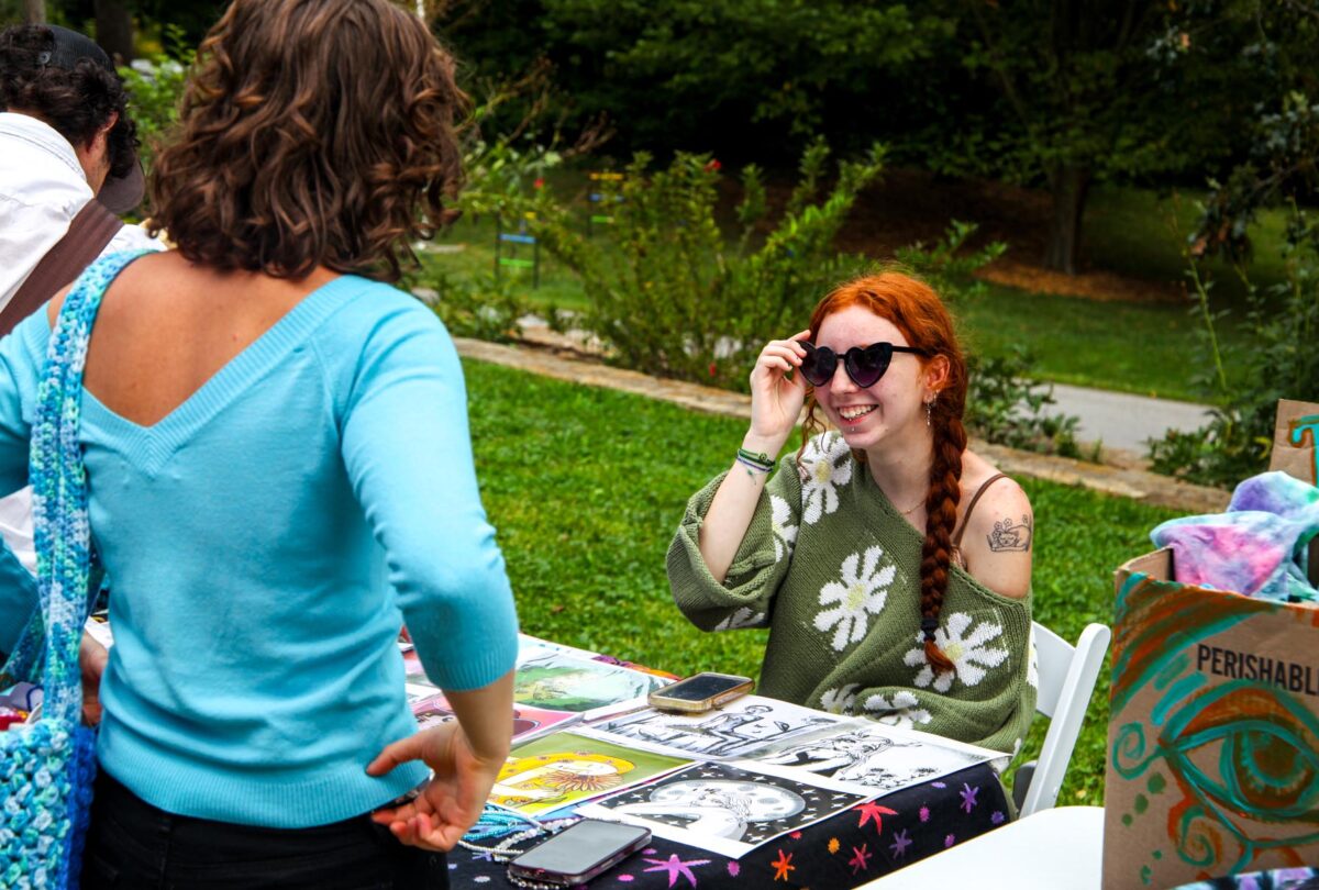 A student smiles from behind a table of their art work they are selling at a craft fair.