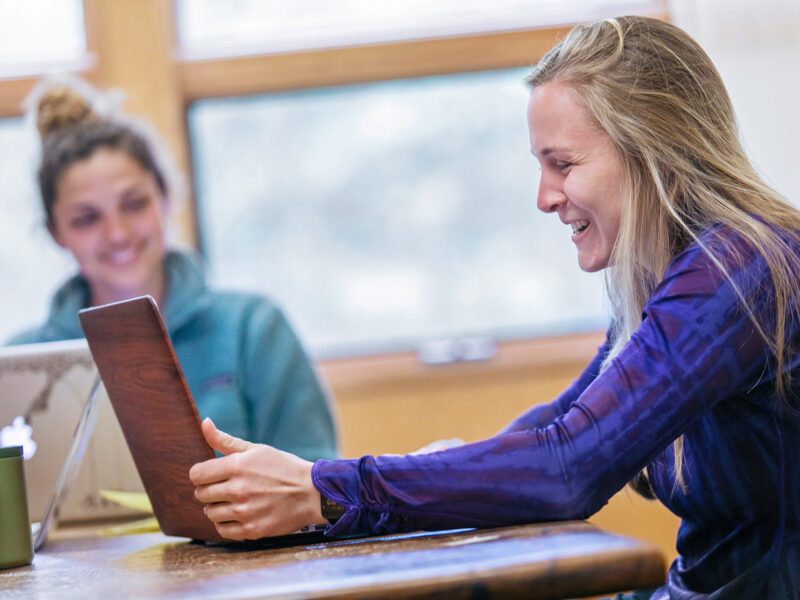 Two students sit beside each other looking at their laptops smiling