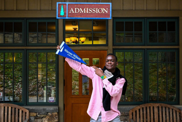 Student Munashe stands smiling and holding a warren wilson pennant in front of the admissions office.