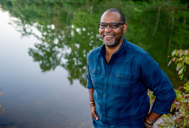 J. Drew Lanham stands by a body of water, which reflects trees behind him. He is wearing glasses and a blue button-down shirt with his sleeves rolled up.