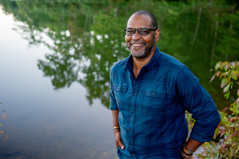 J. Drew Lanham stands by a body of water, which reflects trees behind him. He is wearing glasses and a blue button-down shirt with his sleeves rolled up.