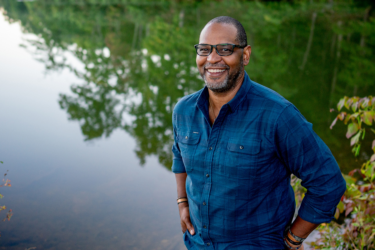 J. Drew Lanham stands by a body of water, which reflects trees behind him. He is wearing glasses and a blue button-down shirt with his sleeves rolled up.