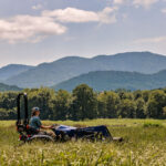 A student drives an Electric Tractor through a pasture with a beautiful mountain range in the background.