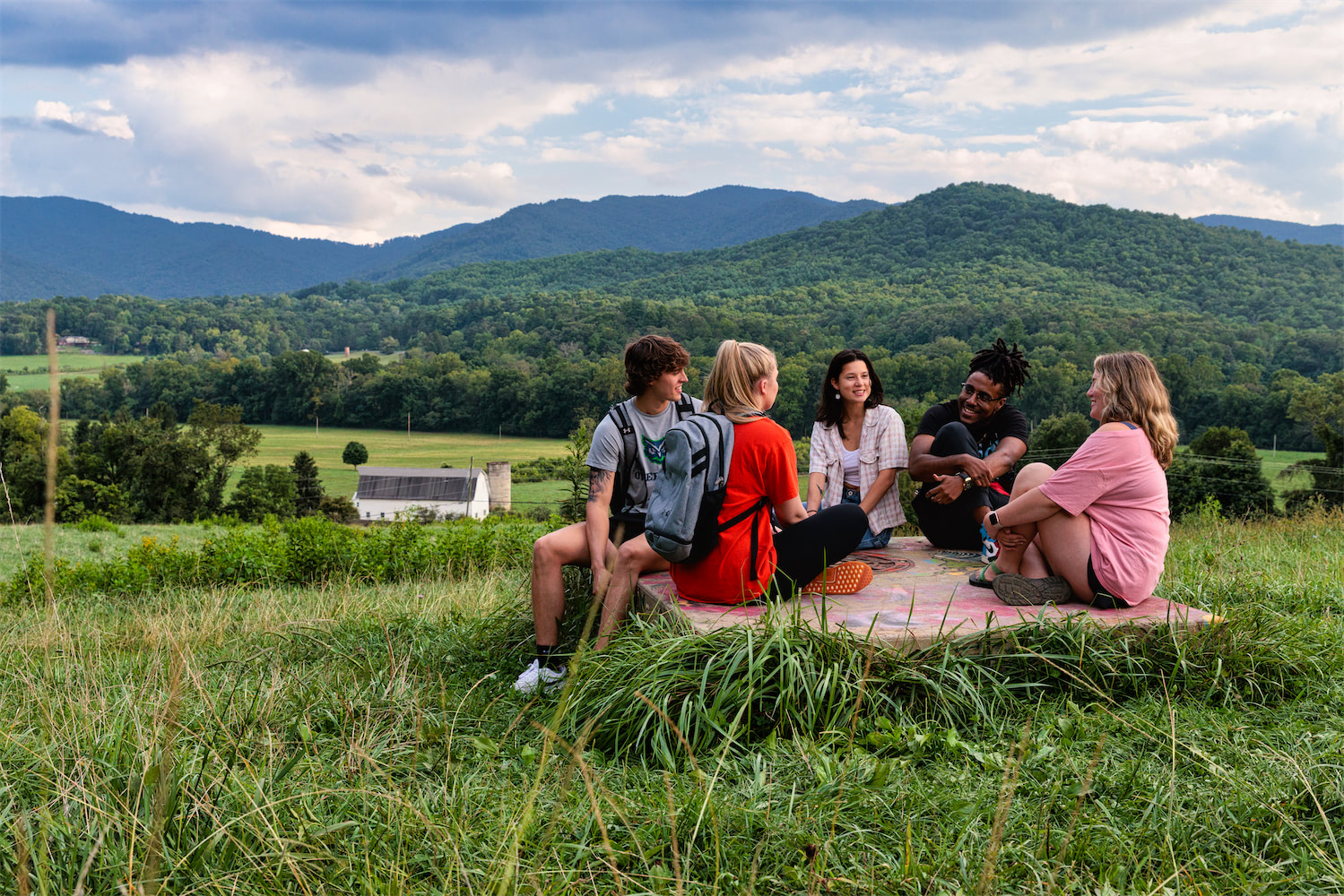 A group of students sit on a concrete pad smiling and laughing. Behind them is a beautiful mountain vista and the white barn.