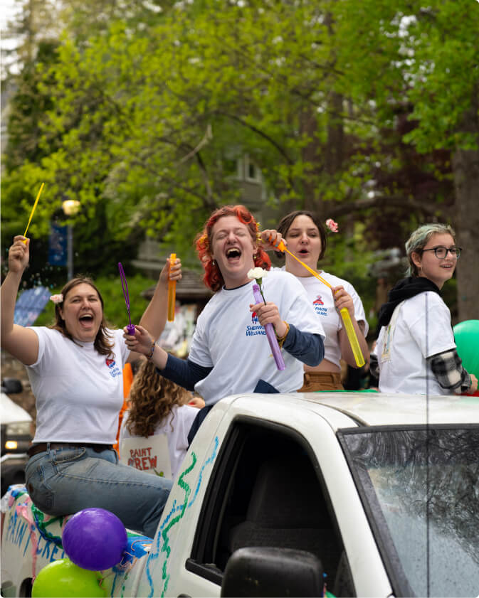 A group of students sit and crouch in the back of a pickup truck that is a parade float in the Big Gay Earth Day Parade. The students are blowing bubbles, smiling, and cheering.