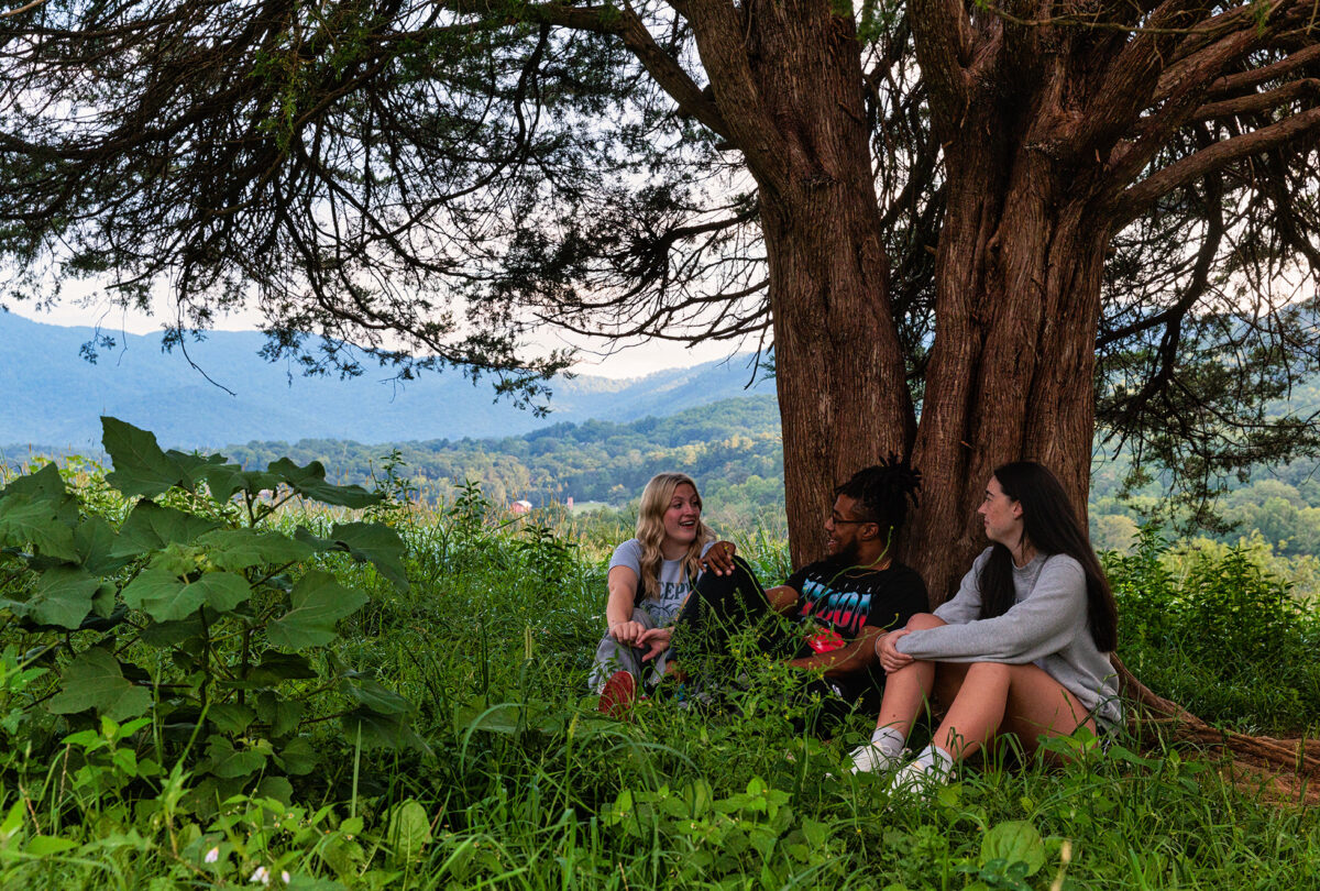 Students sit under a tree in Dogwood pasture. A beautiful view of the valley is their backdrop.