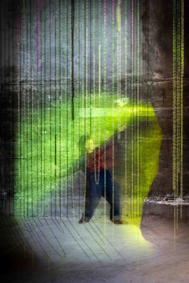 Professor Kathryn Cellerini Moore poses at an exhibition behind metal chains that hang from the ceiling. A bright green and yellow spotlight shines towards them.