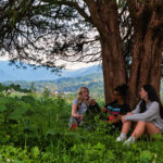 Students sit under a tree in Dogwood pasture. A beautiful view of the valley is their backdrop.